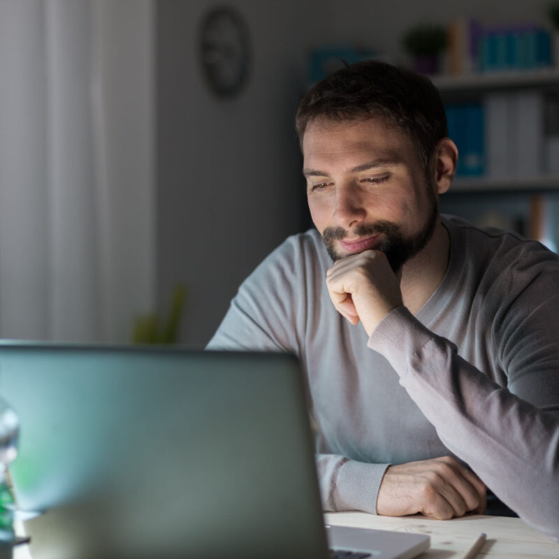 Young smiling man with hand on chin connecting to internet and using a laptop