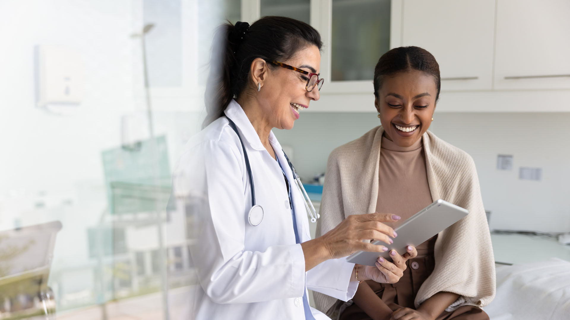 Happy senior practitioner woman and cheerful young African patient woman watching examination medical report on tablet computer, using digital technology